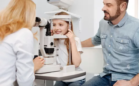 A young girl having her eyes examined by an optometric while her father watches along.